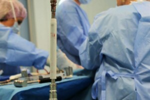 Operāciju telpa ar ķirurgiem, medicīnas instrumentiem un asins traipiem uz galda. Redzama operācijas ierīce.