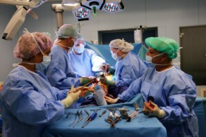 Ķirurģiska operācija, kurā piedalās pieci mediķi, izmantojot medicīniskos instrumentus un aprīkojumu.