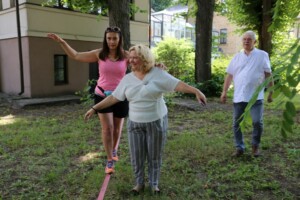 Divas sievietes staigā pa šauru virvi, vīrietis stāv fonā. Rīgas 1. slimnīca, rehabilitācija un līdzsvara vingrinājumi.