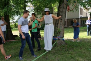 Cilvēki piedalās aktivitātē, balansējot uz virves zem kokiem Rīgas 1. slimnīcas teritorijā.