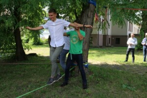 Divi vīrieši balansē uz virves, viens palīdz otram. Fons: Rīgas 1. slimnīcas teritorija ar skatītājiem.