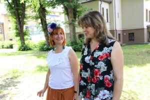 Divas sievietes staigā pa zaļu zāli, viena ar ziedu vainagu, otra ar ziedu apdrukas blūzi. Rīgas 1. slimnīca.