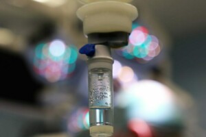 Infūzijas sistēma ar šķidrumu, fons ar medicīnas personālu un operācijas apgaismojumu. Rīgas 1. slimnīca.