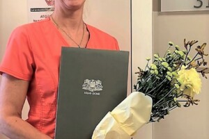 Sieviete rozā medicīnas tērpā ar ziediem un dokumentu, stāv pie Rīgas 1. slimnīcas ambulances.