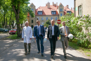 Četri vīrieši, tostarp ārsts, staigā pa Rīgas 1. slimnīcas teritoriju, nēsājot ķiveres. Fons ar ēku un kokiem.