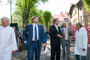 Grupa vīriešu, tostarp ārsti un amatpersonas, staigā pa ielu pie Rīgas 1. slimnīcas. Redzami koki un ēkas.