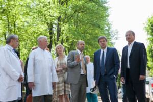 Grupa cilvēku, tostarp mediķi un vīrieši uzvalkos, diskusijā Rīgas 1. slimnīcas teritorijā. Zaļš fons.