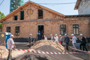 Būvniecības vieta pie Rīgas 1. slimnīcas, cilvēki ar ķiverēm, smilšu un grants kaudzes.
