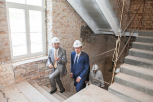 Divi vīrieši ar ķiverēm kāpj pa kāpnēm, Rīgas 1. slimnīcas renovācijas laikā. Redzami akmens mūri un logi.