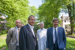 Četri vīrieši stāv parkā, zaļās kokā, Rīgas 1. slimnīcas apkārtnē. Viens ģērbies baltā medicīnas tērpā.