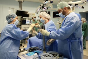 Četri medicīnas darbinieki operāciju zālē strādā ar instrumentiem un aprīkojumu. Redzami medicīnas piederumi.