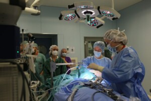 Operāciju telpa ar medicīnas personālu, kas veic operāciju. Redzami instrumenti un medicīnas aprīkojums.