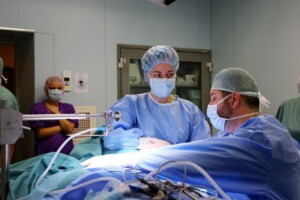 Ārsti operācijas laikā Rīgas 1. slimnīcā, valkājot medicīniskās drēbes un maskas. Fons: operāciju zāle.