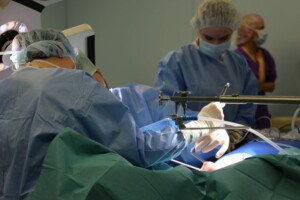 Divi ķirurgi veic operāciju, tērpti medicīniskās drēbēs un maskās, ar instrumentiem rokās.