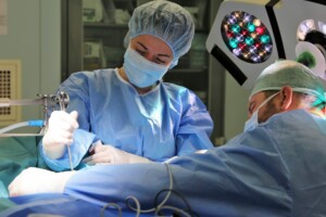 Divi ķirurgi veic operāciju, tērpti zilos medicīnas tērpos un maskās, izmantojot medicīniskos instrumentus.