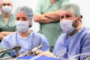 Divi medicīnas darbinieki operāciju zālē, valkājot maskas un zilu apģērbu, koncentrējas uz procedūru.