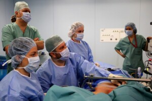 Četri medicīnas darbinieki operācijas telpā, valkājot maskas un medicīnisko apģērbu. Redzama operācijas gulta.