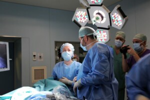 Ārsti operāciju zālē, runājot un gatavojoties procedūrai. Redzams monitors un operāciju gaismas.