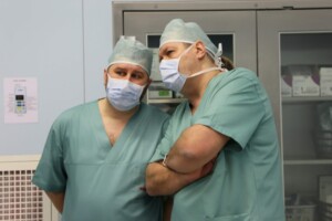 Divi medicīnas darbinieki zaļās drēbēs un maskās apspriežas slimnīcas telpā. Fons ar medicīnas aprīkojumu.