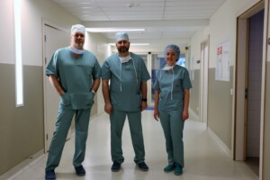 Trīs medicīnas darbinieki zaļās drēbēs stāv slimnīcas gaitenī. Rīgas 1. slimnīca.