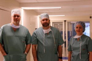 Trīs mediķi zaļās drēbēs un cepurēs stāv slimnīcas koridorā. Rīgas 1. slimnīca.