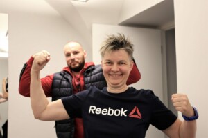 Divi cilvēki pozē, rādot muskuļus. Sieviete valkā T-kreklu ar uzrakstu "Reebok". Fons ir gaišs un neitrāls.