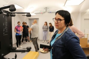 Rīgas 1. slimnīcas rehabilitācijas telpa ar cilvēkiem un KINESIS aprīkojumu. Sieviete ar telefonu rokās.