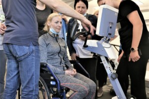 Attēlā redzama sieviete ratiņkrēslā, kas saņem instrukcijas par rehabilitācijas iekārtu. Apkārt ir cilvēki.