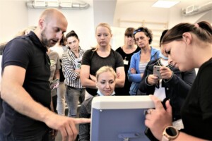 Grupa cilvēku, kas vēro instrukcijas, kā izmantot ierīci Rīgas 1. slimnīcā. Redzams ekrāns un rokas.