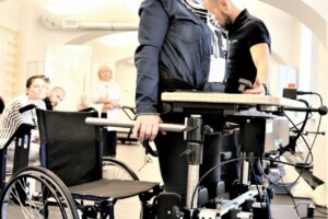 Sieviete stāv uz rehabilitācijas ierīces, blakus ratiņkrēslam, ar speciālistu, Rīgas 1. slimnīca.