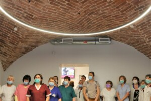 Rīgas 1. slimnīcas darbinieki grupā, tērpti medicīniskajās drēbēs un maskās, stāv telpā ar ķieģeļu griestiem.