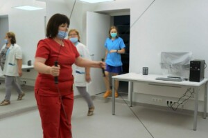 Sieviete sarkanā apģērbā izmanto medicīnisko aprīkojumu Rīgas 1. slimnīcā. Fons ir klīniska vide.