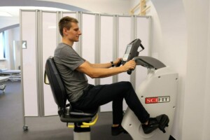 Vīrietis vingro uz SCI FIT trenažiera Rīgas 1. slimnīcā, fona ekrāni nodrošina privātumu.
