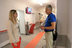 Divi cilvēki sarunājas Rīgas 1. slimnīcas gaidīšanas zonā, redzams ekrāns ar numuriem.
