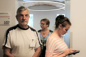 Vīrietis un divas sievietes gaida pie numura 107 Rīgas 1. slimnīcā, sieviete lieto planšetdatoru.