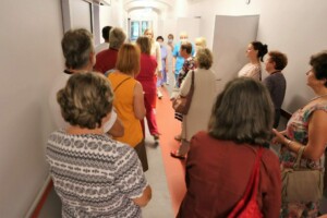 Grupa cilvēku gaida koridorā, daži medicīnas darbinieki ir redzami attālumā. Rīgas 1. slimnīca.