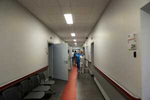Gaišs slimnīcas gaiteņš ar numuru zīmēm, mediķi un gaidīšanas krēsli. Numurs 224, stāvs 7.