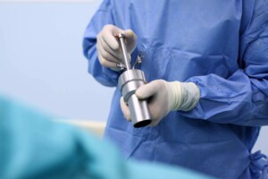 Ārsts, tērpies zilos medicīnas tērpos, tur ķirurģisku instrumentu. Fons ir gaiši zils.