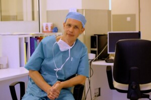 Ārsts ar zilu medicīnisko apģērbu sēž pie galda Rīgas 1. slimnīcā, fonā dators un dokumenti.