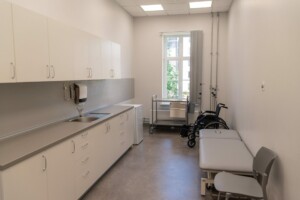 Gaiša medicīnas telpa ar galdu, krēslu, ratiņkrēslu un logu, kas sniedz skatu uz ārpusi.