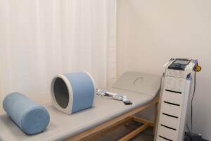 Fizioterapijas telpa ar ārstniecības galdu, zilu rullīti un medicīnas iekārtu. Rīgas 1. slimnīca.