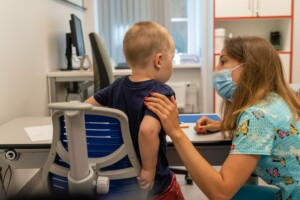 Bērns sēž pie galda, medicīnas darbinieks runā ar viņu, abi valkā maskas. Rīgas 1. slimnīcas telpa.
