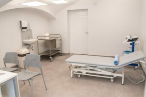 Tīra medicīnas telpa ar izmeklēšanas galdu, krēsliem un medicīnas aprīkojumu. Rīgas 1. slimnīca.