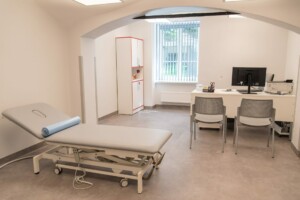Gaiša medicīnas telpa ar izmeklēšanas galdu, datoru un divām krēslām. Logi ar žalūzijām.
