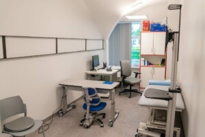 Ārsta kabinets ar galdu, krēsliem un datoru, gaišs un sakārtots, Rīgas 1. slimnīcā.