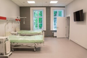 Gultas ar zaļām gultasveļām Rīgas 1. slimnīcas telpā, logi ar aizkariem un televizors pie sienas.