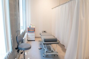 Rīgas 1. slimnīcas telpa ar ārstniecības gultu, krēslu un aizkaru, gaiša un sakopta vide.
