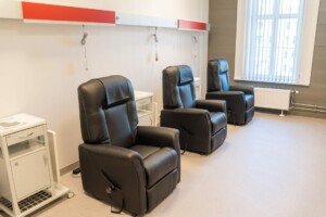 Trīs melni atpūtas krēsli Rīgas 1. slimnīcas telpā, blakus baltām mēbelēm un logiem ar žalūzijām.