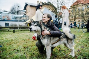 Sieviete apskauj suni, fonā redzamas citas personas un Rīgas 1. slimnīcas ēka. Zāle un telts.