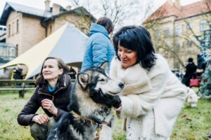 Divas sievietes ar suni, viens no viņām tiek glāstīts. Fons: pasākums ārpus telpām ar cilvēkiem un teltīm.